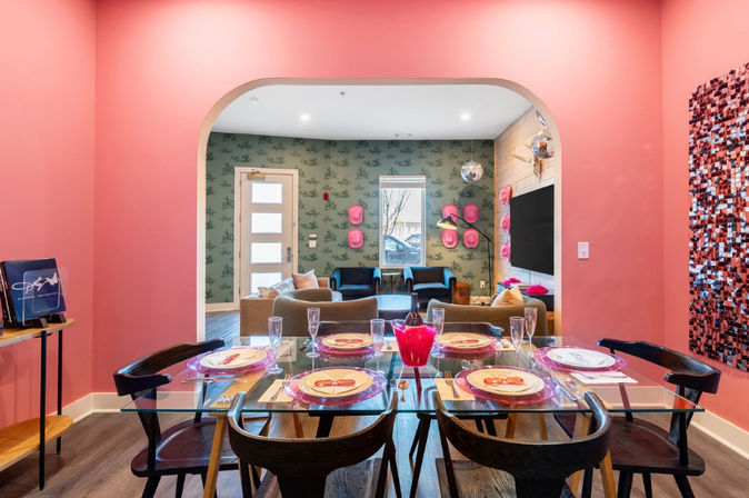 Cheerful pink dining area with glass table set for six, opening through an arched doorway to a modern apartment lounge with green patterned wallpaper, cozy seating and disco-ball accents.