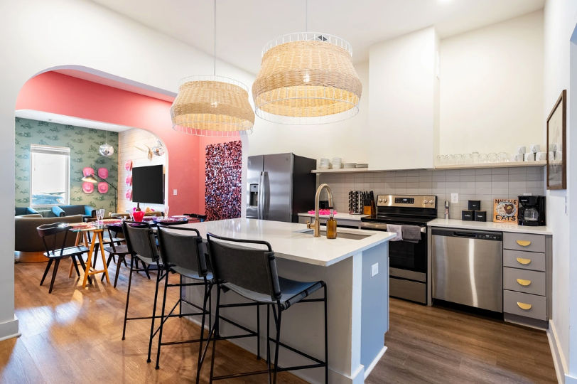 Bright modern open-concept kitchen with white island and black bar stools, woven pendant lights, stainless steel appliances, playful pink arched accent wall and adjacent cozy living area on hardwood floors.