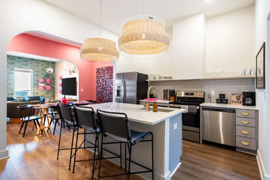 Bright modern open-concept kitchen with white island and black bar stools, woven pendant lights, stainless steel appliances, playful pink arched accent wall and adjacent cozy living area on hardwood floors.