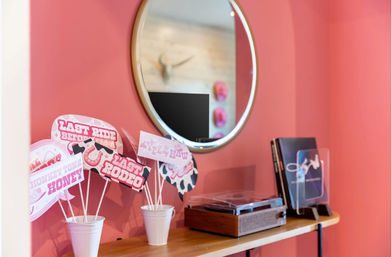 Pink-walled vignette with a round mirror above a wooden shelf holding a retro turntable, vinyl sleeves, and white cups of playful rodeo-themed photo props (’Last Rodeo’)