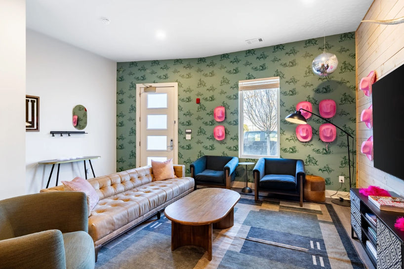 Bright modern living room with tufted tan leather sofa, wooden coffee table, two blue armchairs, green patterned wallpaper accented by playful pink hat wall decor and a hanging disco ball, rug, TV and a window showing a tree outside.