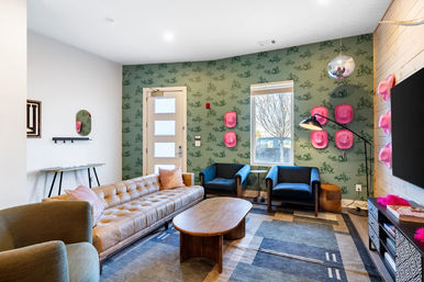 Bright modern living room with tufted tan leather sofa, wooden coffee table, two blue armchairs, green patterned wallpaper accented by playful pink hat wall decor and a hanging disco ball, rug, TV and a window showing a tree outside.