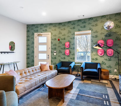 Bright modern living room with tufted tan leather sofa, wooden coffee table, two blue armchairs, green patterned wallpaper accented by playful pink hat wall decor and a hanging disco ball, rug, TV and a window showing a tree outside.