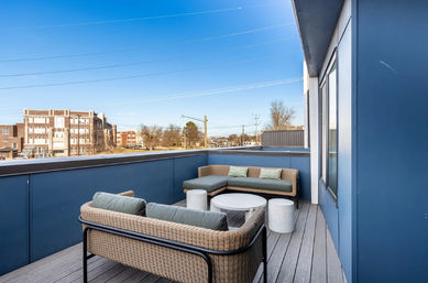 Modern urban rooftop balcony with wicker sectional sofa and green cushions, round white coffee tables, blue privacy walls and downtown buildings in the distance under a clear blue sky