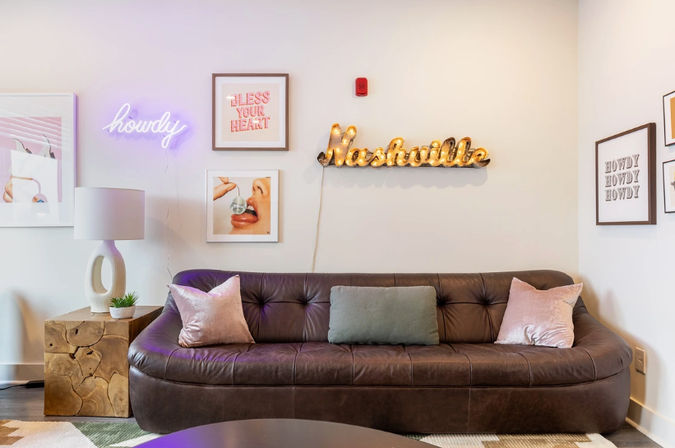 Cozy Nashville-themed lounge with a curved brown leather sofa, velvet pastel pillows, wood block side table and lamp, neon 'howdy' sign and lit marquee 'Nashville' among framed wall art.