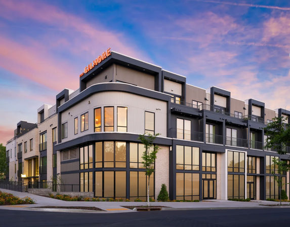 Modern three-story mixed-use building on an urban corner with a curved glass facade, balconies and rooftop sign, warm interior lights and landscaping beneath a pink-purple sunset sky.