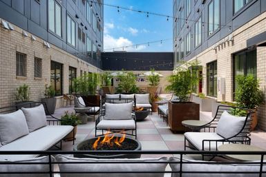 Modern urban courtyard patio between two mid-rise buildings with cushioned metal lounge chairs, low round fire pits, potted plants, and string lights overhead under a blue sky