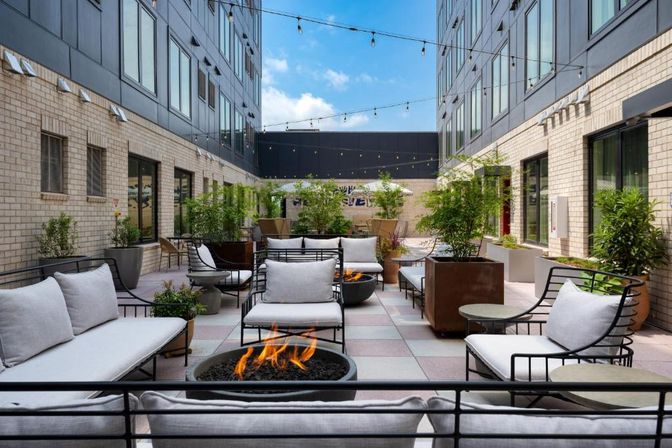 Modern urban courtyard patio between two mid-rise buildings with cushioned metal lounge chairs, low round fire pits, potted plants, and string lights overhead under a blue sky