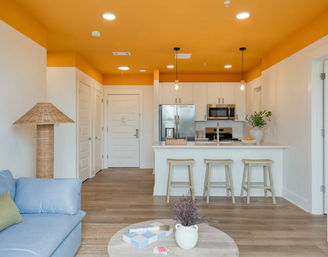 Modern open-concept apartment interior with vibrant orange ceiling, white kitchen cabinets, stainless steel fridge and microwave, breakfast bar with three wooden stools, pendant lights, light wood floors, blue sofa, wicker floor lamp and round coffee table with vase