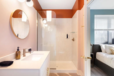 Chic blush-pink bathroom with glass walk-in shower, round brass mirror, white vanity with gold faucet and amber soap dispenser, open door to teal-accent bedroom