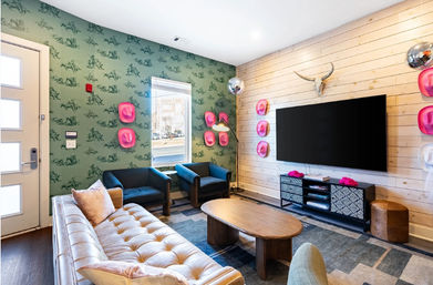 Cozy eclectic lounge with a tufted tan leather sofa, two navy armchairs, wooden coffee table and large wall-mounted flat-screen TV on light shiplap, green patterned wallpaper, decorative pink cowboy hats and disco balls.