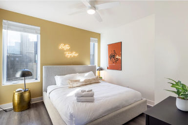 Sunlit modern city apartment bedroom with a textured gray upholstered bed, crisp white linens and folded towels, mustard accent wall featuring a neon "Better Together" sign, two tall windows with city view, gold side table with black lamp, orange framed art, ceiling fan and potted plant.