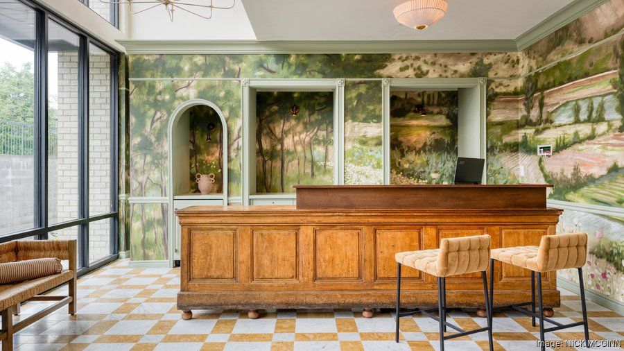 Boutique hotel lobby reception with a long wooden check-in desk, tufted tan bar stools, scenic green landscape mural walls, checkerboard tile floor and floor-to-ceiling windows.