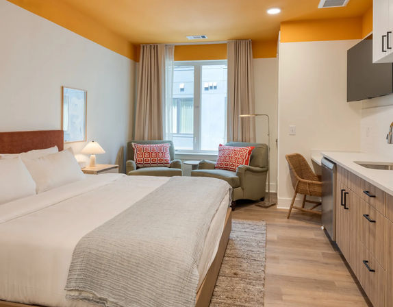 Bright, cozy studio hotel room with a king-size bed in white linens, gray throw, two armchairs with red patterned pillows by a sunlit window, kitchenette with wood cabinets and wicker chair, warm yellow ceiling accent.