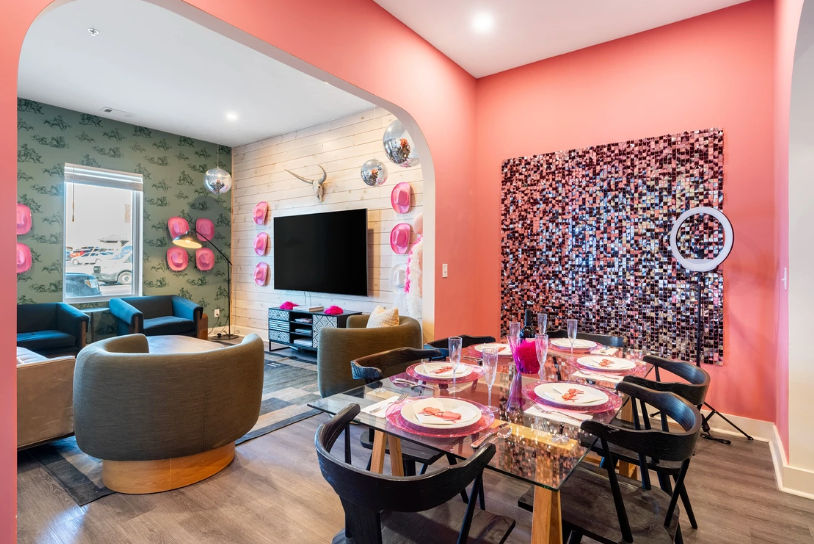 Vibrant coral-pink open-plan apartment lounge and dining area with glass table set for six, mosaic wall art, round gray swivel chairs, teal sofas, and TV on a shiplap wall.