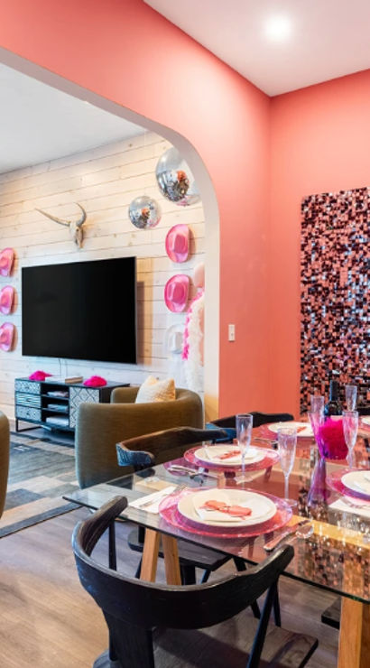 Vibrant coral-pink open-plan apartment lounge and dining area with glass table set for six, mosaic wall art, round gray swivel chairs, teal sofas, and TV on a shiplap wall.