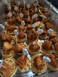 Aluminum catering tray of mini chicken-and-waffle appetizers: crispy fried chicken bites on mini round waffles, each secured with a wooden skewer topped with a white snowflake pick.