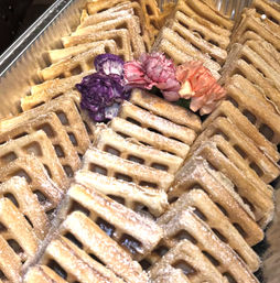 Powdered-sugar dusted square waffles neatly stacked in a metal tray, garnished with purple, pink and peach carnation flowers.