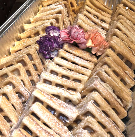 Powdered-sugar dusted square waffles neatly stacked in a metal tray, garnished with purple, pink and peach carnation flowers.