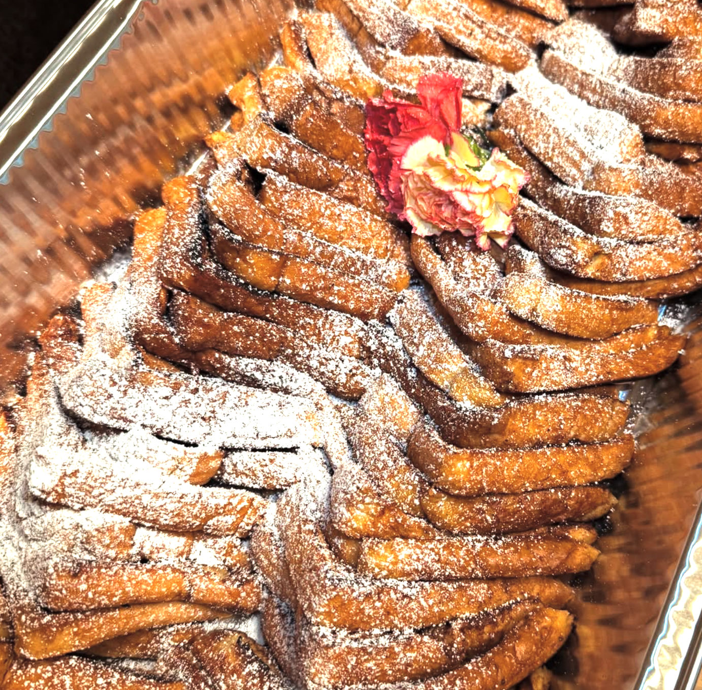 Catering tray of golden twisted fried pastries dusted with powdered sugar, garnished with a pink carnation
