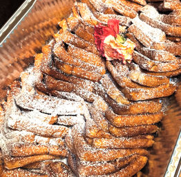 Catering tray of golden twisted fried pastries dusted with powdered sugar, garnished with a pink carnation