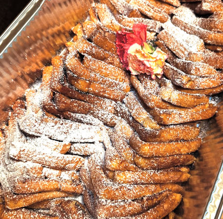 Catering tray of golden twisted fried pastries dusted with powdered sugar, garnished with a pink carnation