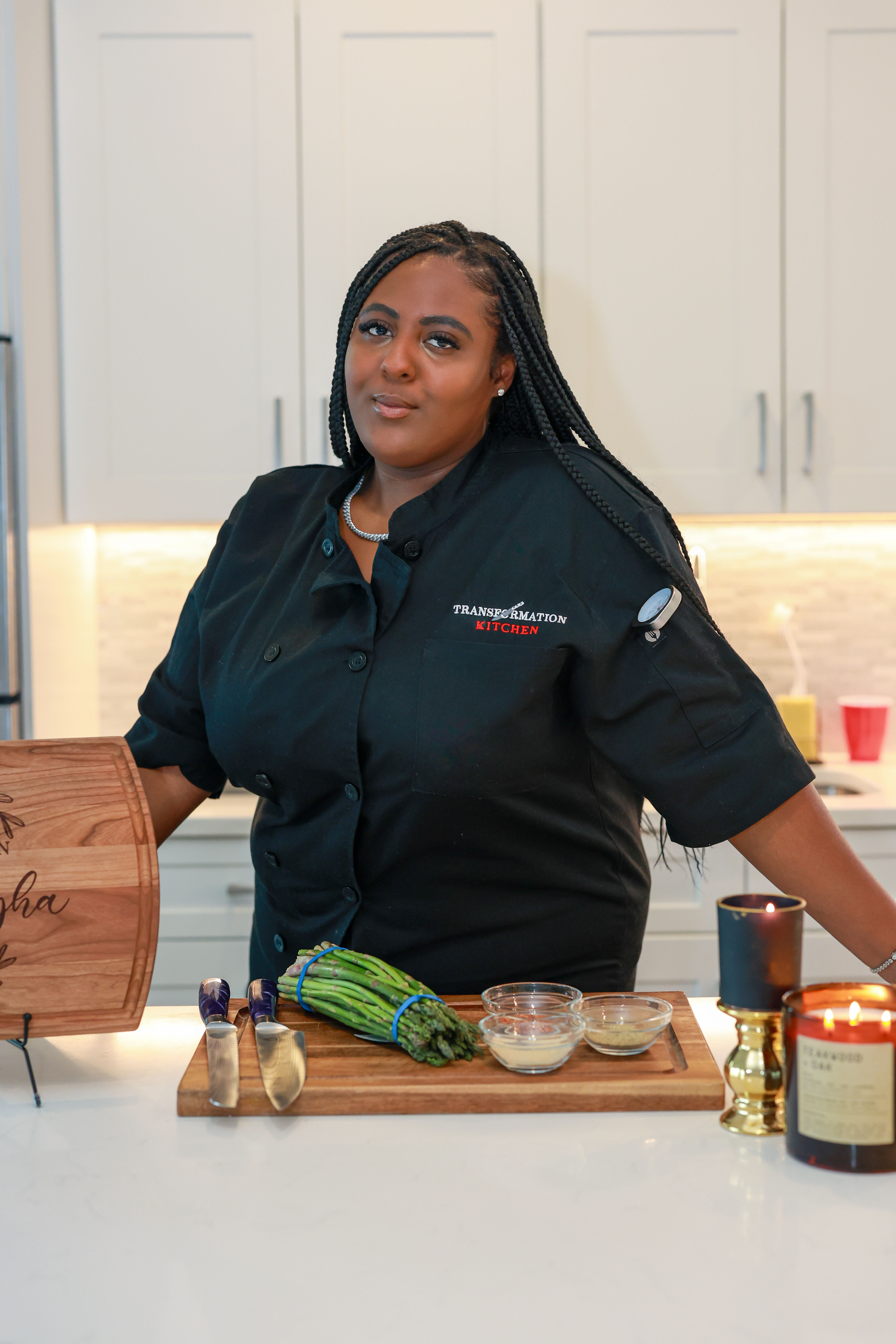 Chef in a black coat in a modern white kitchen preparing asparagus on a wooden cutting board with knives and small glass seasoning bowls.