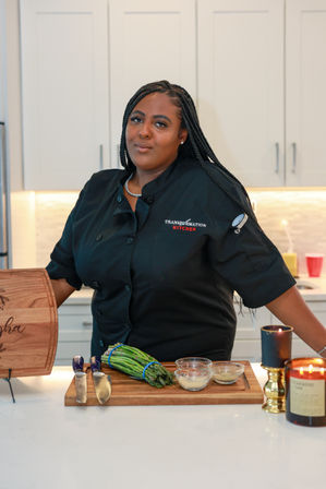 Chef in a black coat in a modern white kitchen preparing asparagus on a wooden cutting board with knives and small glass seasoning bowls.