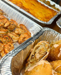 Catering aluminum trays with seasoned grilled shrimp, a cheesy baked casserole, and crispy stuffed fried rolls with savory filling drizzled with sauce — party-style takeout platter