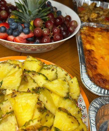 Bright party spread featuring a plate of fresh pineapple chunks, a bowl of grapes, strawberries and blueberries, and a cheesy baked casserole in foil pans on a wooden table