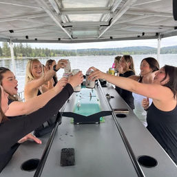 Group of friends cheerfully toasting canned drinks on a covered pontoon boat during a lake outing, calm water and forested shoreline in the background