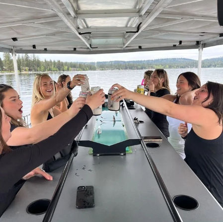 Group of friends cheerfully toasting canned drinks on a covered pontoon boat during a lake outing, calm water and forested shoreline in the background