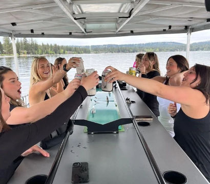 Group of friends cheerfully toasting canned drinks on a covered pontoon boat during a lake outing, calm water and forested shoreline in the background