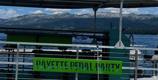 Bright green sign on a covered pedal-party boat at a scenic mountain lake, calm blue water, forested shoreline and rocky peaks under a partly cloudy sky.