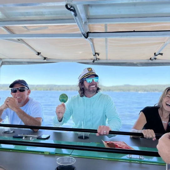 Smiling group on a pontoon boat during a sunny lake outing, person in a captain's hat shaking a maraca.