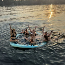 Six friends in swimsuits wearing captain hats on a round inflatable raft in a forest-lined lake at sunset, holding drinks and cheering.
