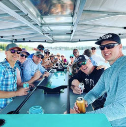 Smiling group of friends on a covered pontoon boat on a sunny lake, holding canned drinks and enjoying a casual summer lake cruise