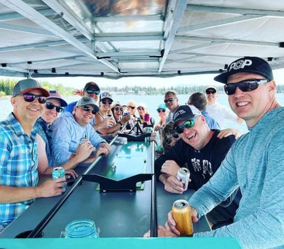 Smiling group of friends on a covered pontoon boat on a sunny lake, holding canned drinks and enjoying a casual summer lake cruise