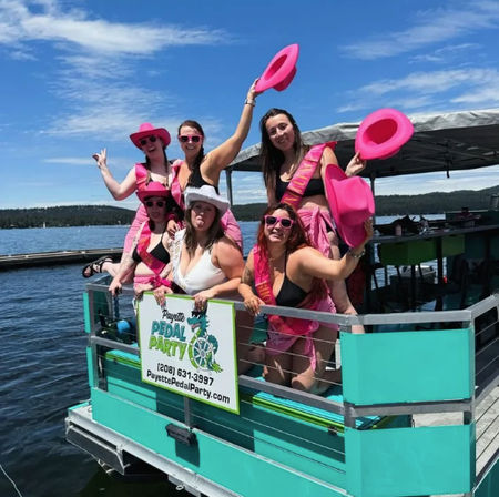 Six women in pink hats, sunglasses and sashes celebrating on a turquoise pedal party boat at a sunny lake, waving cowboy hats and posing for a bachelorette-style outing