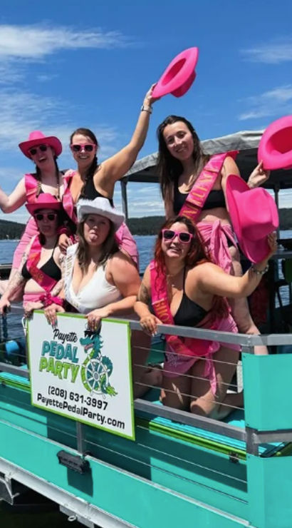 Six women in pink hats, sunglasses and sashes celebrating on a turquoise pedal party boat at a sunny lake, waving cowboy hats and posing for a bachelorette-style outing