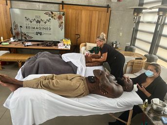 Two men unwind on massage tables while therapists provide spa treatments in a bright indoor wellness-retreat space with decorative signage in the background.
