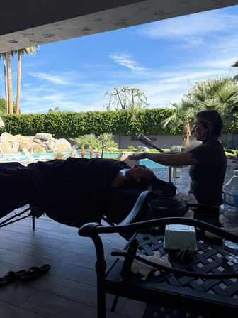Poolside facial on a shaded patio — a therapist uses a steamer over a client on a treatment table with a sparkling pool, palm trees and bright blue sky in the background.