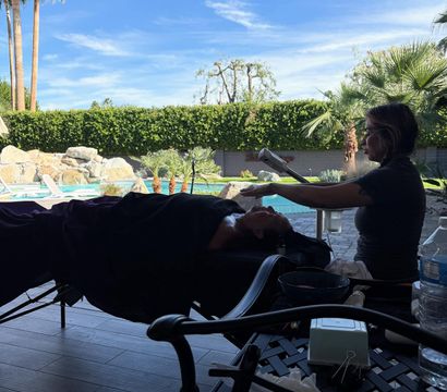 Poolside facial on a shaded patio — a therapist uses a steamer over a client on a treatment table with a sparkling pool, palm trees and bright blue sky in the background.