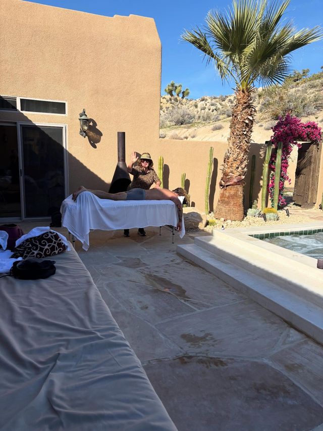 Sunlit desert patio spa scene with a massage table by a pool, therapist waving next to a person receiving a massage, palm tree, tall cacti, stucco wall and rocky hillside in the background