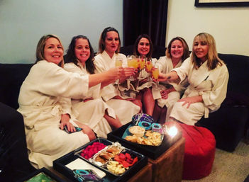 Six women in white spa robes sitting on a couch in a cozy living room, toasting with mimosas over trays of snacks and appetizers — girls' night spa party.