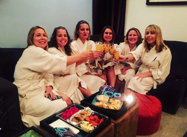 Six women in white spa robes sitting on a couch in a cozy living room, toasting with mimosas over trays of snacks and appetizers — girls' night spa party.