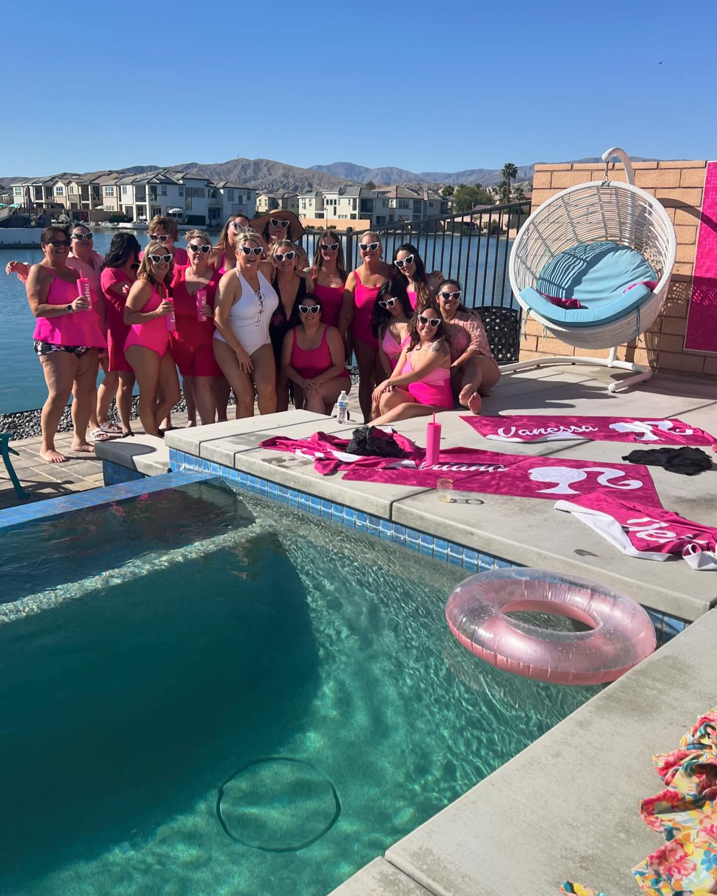Group of women in matching pink swimsuits and heart-shaped sunglasses at a sunny lakeside backyard pool party with pink towels, inflatable ring, hanging wicker chair, and mountain views