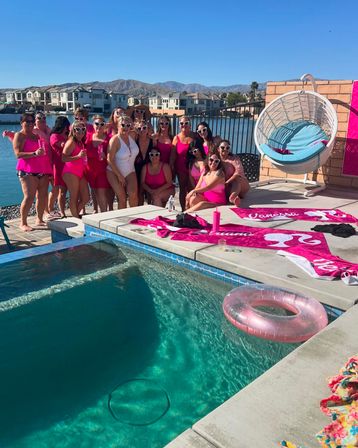 Group of women in matching pink swimsuits and heart-shaped sunglasses at a sunny lakeside backyard pool party with pink towels, inflatable ring, hanging wicker chair, and mountain views