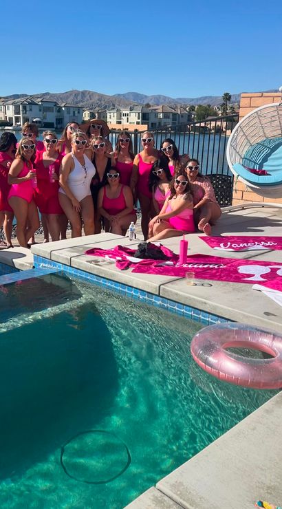 Group of women in matching pink swimsuits and heart-shaped sunglasses at a sunny lakeside backyard pool party with pink towels, inflatable ring, hanging wicker chair, and mountain views