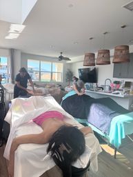 Sunlit at-home massage party in a residential living room — multiple people lie face-down on portable massage tables while therapists give massages, with large windows showing neighboring houses and distant mountains.